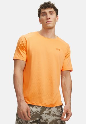 Jeune homme portant un t-shirt de sport à manches courtes orange vif et un short camouflage gris, debout devant un fond clair uni.