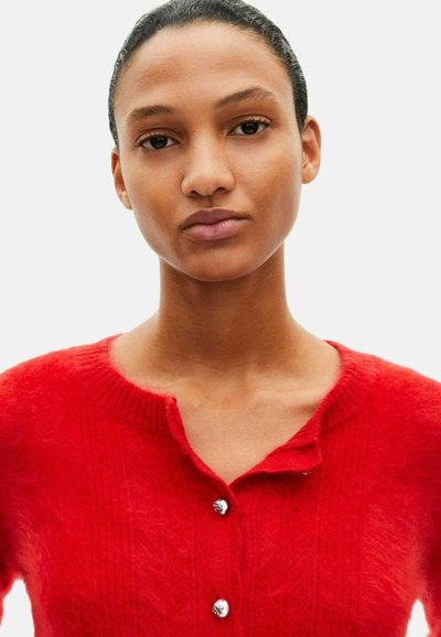 Rød cardigan lavet af blødt, tekstureret stof med rund halsudskæring, små knapper og lodrette ribstrikmønstre.