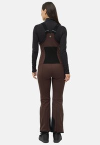 Kvinde iført brune bibbukser med sort elastisk rygpanel og sorte justerbare stropper over en sort langærmet top, stående med ryggen til.