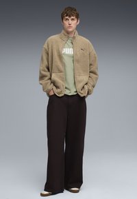 Beige fleecejakke med høj krave, lagdelt over en lysegrøn t-shirt med et iøjnefaldende hvidt logo. Sammen med brede sorte bukser og sneakers.
