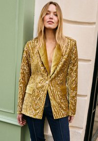 Gouden blazer met pailletten, een fluwelen notch-revers, een gestructureerd patroon en twee voorzakken, gecombineerd met donkere spijkerbroek.