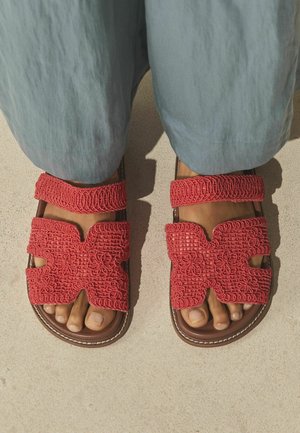 Füße, die rote gewebte Sandalen mit Blumenmustern und zwei Riemen tragen, kombiniert mit hellblauen Hosen, stehend auf einer hellen Oberfläche.