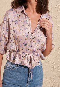 Blouse florale violette clair en satin, avec un col, des manches courtes et une taille cintrée avec un détail à nouer. Portée avec un short en denim.