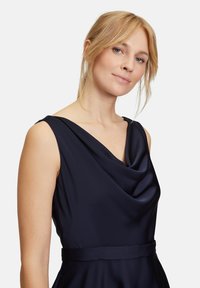 Robe sans manches bleu marine avec un col drapé, une silhouette épurée et une texture satinée. Comprend une ceinture assortie à la taille.