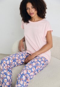 Šviesiai rožinė marškinėlių su trumpomis rankovėmis viršutinė dalis su balta užrašu ir gėlių raštuota mėlyna kelnėmis, puoštomis rožinėmis ir violetinėmis gėlėmis. Minkšta, laisvai krentančio kirpimo.