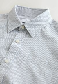 Pelēka un baltā svītraina krekla ar pogām, kas pazīstama ar vērstu apkakli, trim pogām un vienu krūšu kabatu, izgatavota no teksturēta auduma.