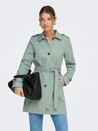 Trench-coat vert clair avec une taille ceinturée, une fermeture à boutons sur le devant et des épaulettes. Le modèle tient un sac à main noir et porte un jean bleu.