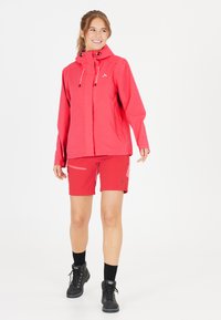 Ljus röd vattentålig jacka med justerbar huva och framzip, tillsammans med matchande röda shorts med ljusare detaljer och sidofickor. Svarta vandringskängor.