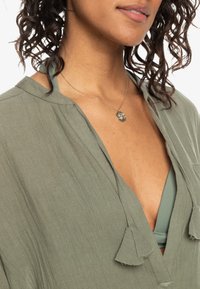 Blusa verde oliva con scollo a V, in tessuto leggero e dettagli con frange. Indossata sopra un top bikini verde chiaro. Capelli ricci visibili.
