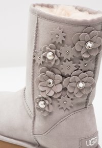 Botte d'hiver en daim gris avec des fleurs en tissu décorées de perles et de strass, montrant une doublure en polaire à l'intérieur.