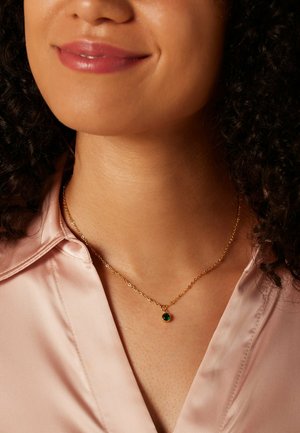 Collier en or avec un petit pendentif en pierre verte, porté avec un blouse en satin légère rose, mettant en valeur le design délicat du collier.