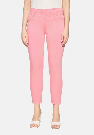 Femme portant un pantalon slim taille haute rose et des sandales blanches à talons carrés ouverts, debout devant un fond uni.
