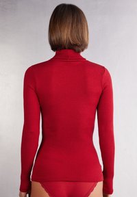Maglietta a collo alto rossa a costine con maniche lunghe, caratterizzata da un design aderente e una texture liscia. La vista posteriore mostra una costruzione semplice e senza cuciture.