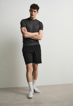 Noor mees seisab käed risti, kandes musta t-särki, musti pükse, valgeid sokke ja valgeid tenniseid ühevärvilise heleda tausta ees.