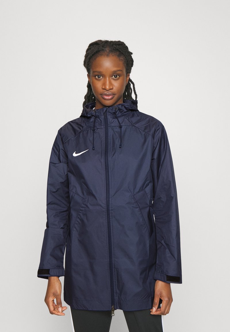 Giacca impermeabile blu navy con cappuccio, zip a tutta lunghezza, tasche laterali e logo Nike bianco sul lato sinistro del petto. Texture liscia, maniche lunghe.
