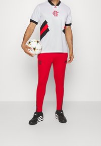 Un maillot de football blanc avec des accents rouges et noirs, associé à des leggings rouges ornés de logos noirs, tenu par une personne avec un ballon de football multicolore.
