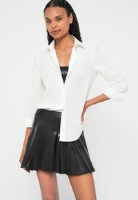 Balta pogājama krekls ar uzruļētām piedurknēm, uzvilkts virs melna tvertnes tipa topa. Valkāts kopā ar melnu, salocītu mini svārku no spīdīga materiāla.