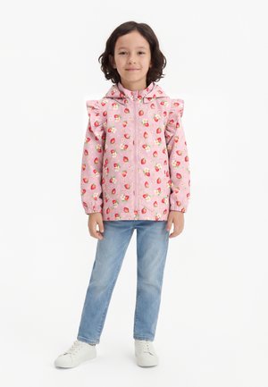 Un niño de pie vestido con una chaqueta rosa con capucha y estampado de fresas y hombros con volantes, jeans azul claro y zapatillas blancas sobre un fondo blanco.
