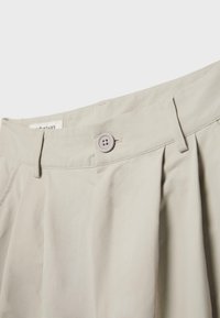 Pantalon beige clair taille haute avec fermeture à boutons, passants pour ceinture et plis sur le devant, présenté sur un fond blanc uni.