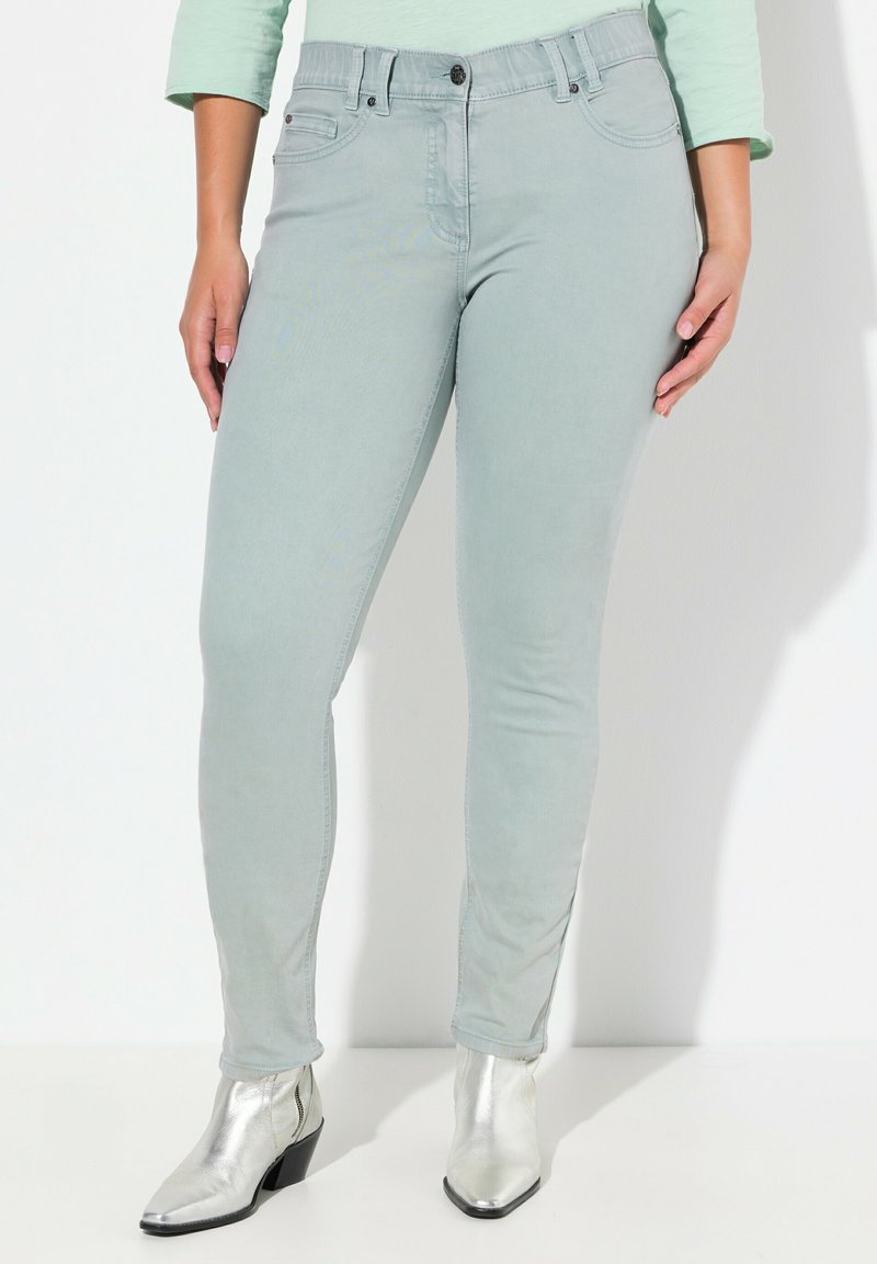 Jeans skinny verde claro con cinco bolsillos, pasadores para cinturón y cierre de botón frontal, combinados con botas de tobillo metálicas plateadas.