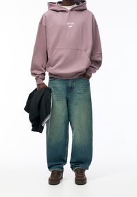 Übergroßer lila Hoodie mit Fronttasche, ausgestattet mit weißen Text. Lockere blaue Jeans aus Denim mit abgewetztem Finish. Schwarze Jacke in der Hand gehalten.
