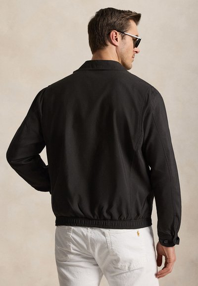 Férfi fekete dzsekiben és fehér nadrágban, hátulról nézve, napszemüveggel és egy kézzel a csípőjén, egyszínű háttér előtt.