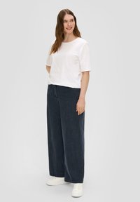 Witte T-shirt met korte mouwen, gecombineerd met donkerblauwe wijde broek. Het model draagt witte schoenen en laat een ontspannen pasvorm en casual stijl zien.