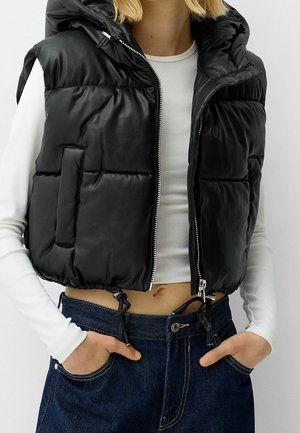 Femme portant une doudoune noire sans manches avec capuche par-dessus un crop top blanc à manches longues, associée à un jean bleu en denim.