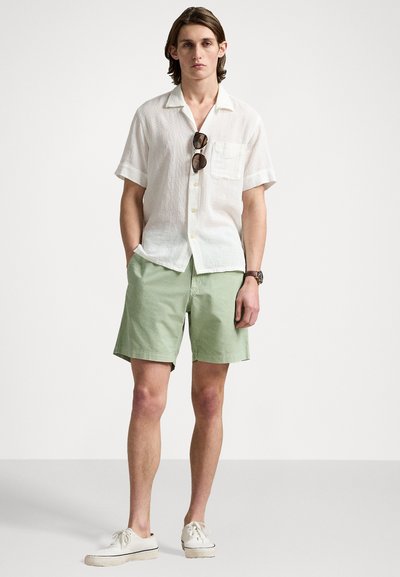 Chemise blanche en lin à manches courtes avec une poche, assortie à un short vert clair. Le modèle porte des lunettes de soleil et des baskets crème.