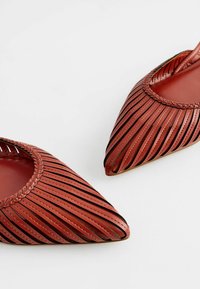 Ballerines en cuir rouge perforé avec bouts pointus et un détail tressé autour de la cheville. Elles présentent des rayures horizontales et une semelle intérieure lisse.