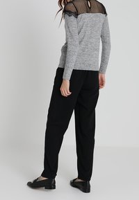 Top grigio a maniche lunghe con schiena in rete nera e rifiniture in pizzo, abbinato a pantaloni neri larghi e scarpe nere basse, visto da dietro.