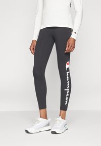 Gambe rivestite con leggings neri Champion con testo del logo sul lato e sneakers atletiche bianche, abbinate a una maglietta bianca a maniche lunghe.