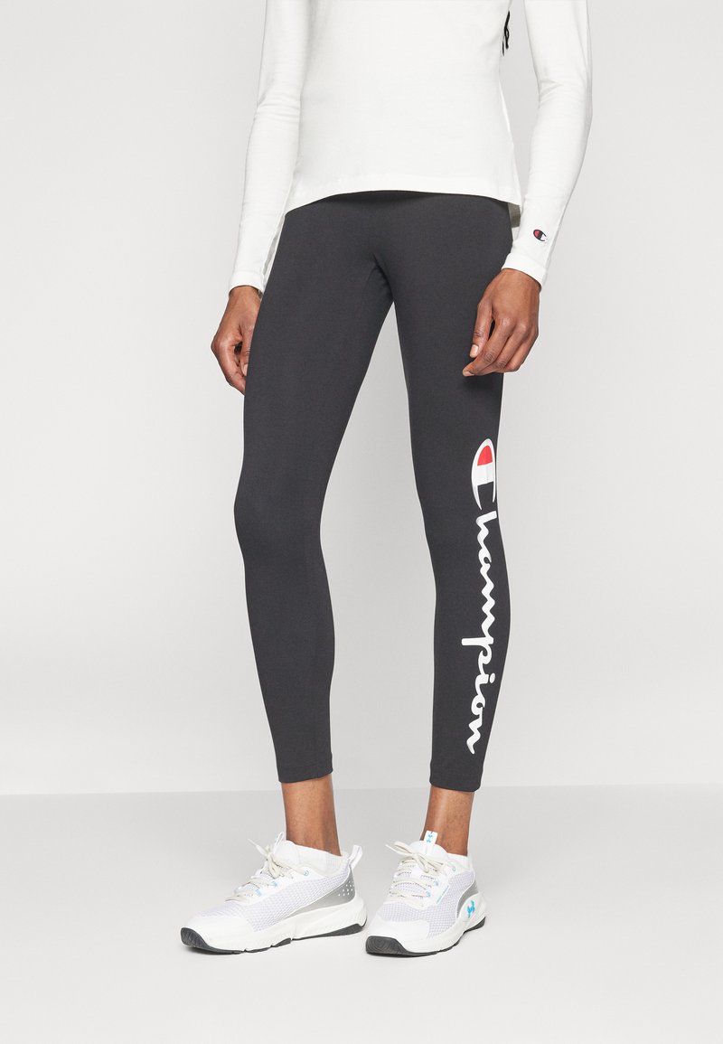 Gambe rivestite con leggings neri Champion con testo del logo sul lato e sneakers atletiche bianche, abbinate a una maglietta bianca a maniche lunghe.