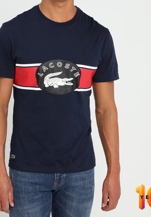 Homme portant un t-shirt Lacoste bleu marine avec une bande rouge et blanche et le logo crocodile, associé à un jean bleu, debout sur fond blanc.