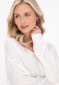 Maglione leggero e trasparente color crema con scollo a V e polsini a costine, abbinato a capelli morbidi e ricci. I dettagli includono trucco minimale e orecchini in argento.