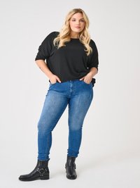 Blondínka stojaca s rukami v vreckách, oblečená v čiernom dlhom rukáve, modrých džínsoch a čiernych členkových topánkach na bielom pozadí.