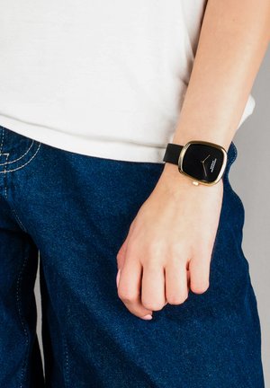 Montre-bracelet avec cadran carré noir et cadre doré portée au poignet gauche, associée à une chemise blanche et un jean en denim bleu.