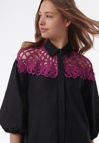 Chemise noire avec un col en dentelle au crochet violet, présentant un design géométrique. La chemise a un devant boutonné et des manches bouffantes.