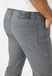 Graue gewebte Hose mit einem Lederpatch mit der Aufschrift "Brax", ausgestattet mit Gesäßtaschen und einem taillierten Schnitt, der ein strukturiertes Stoffdesign zur Geltung bringt.