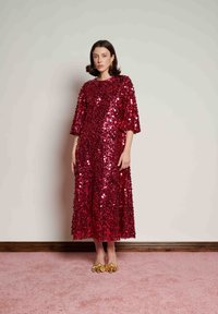 Γυναίκα που στέκεται φορόντας ένα κόκκινο φόρεμα με παγιέτες μήκους έως τη μέση και μανίκια έως τον αγκώνα, καθώς και χρυσά μυτερά παπούτσια, μπροστά σε έναν μονόχρωμο μπεζ τοίχο.