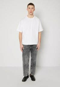 Balta kokvilnas t-krekls ar īsām piedurknēm, aksesuārā pelēki džinsu bikses un melni ādas loferi. Modelis stāv pret neitrālu fonu.