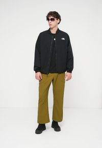 Μαύρο μπουφάν bomber πάνω από μαύρο μπλουζάκι, σε συνδυασμό με χακί πράσινο παντελόνι και μαύρα αθλητικά παπούτσια. Αξεσουάρ περιλαμβάνουν γυαλιά ηλίου.