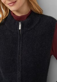 Dunkelgraue, gestrickte ärmellose Weste mit hohem Kragen und silbernem Reißverschluss, getragen über einem maroonfarbenen Langarmshirt. Weicher Stoff.