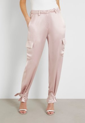 Mujer lleva pantalones cargo de satén rosa claro con bolsillos laterales y puños atados, combinados con sandalias blancas de tacón alto y una camiseta blanca.