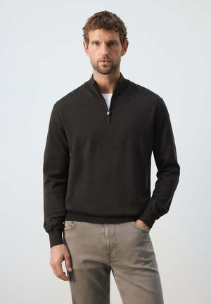Hombre de pie con expresión seria, vistiendo un suéter oscuro con cremallera y pantalones de color claro sobre un fondo liso.