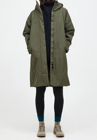 Olijfgroen waterdichte jas, met capuchon, drukknoopsluiting aan de voorkant, relaxed fit, gecombineerd met zwarte leggings en neutrale schoenen.