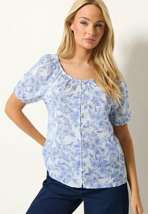 Vrouw met lang blond haar die een witte blouse met een blauw bloemenpatroon en knopen draagt, gecombineerd met een donkerblauwe broek.