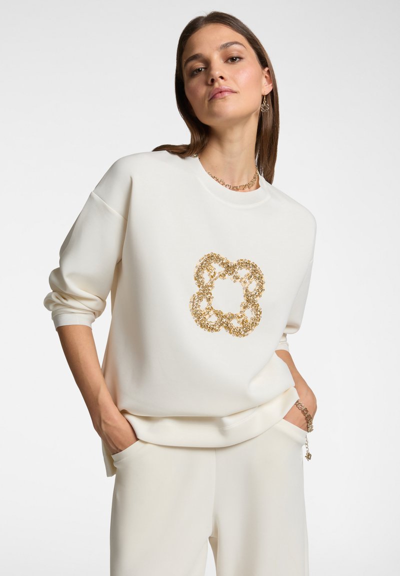 Felpa bianca con un motivo floreale in paillettes dorate testurizzate sul davanti. Presenta un colletto rotondo e una vestibilità rilassata con maniche lunghe.
