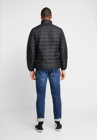 Sort pufferjakke med vandret quiltet tekstur, høj krave, elastiske manchetter, parret med blå jeans og sorte sneakers.