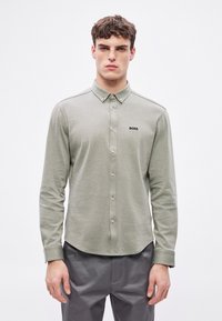 Svetlo zelena srajca s teksturirano tkanino in ovratnikom, z črnim logotipom "BOSS" na prsih in dolgimi rokavi.